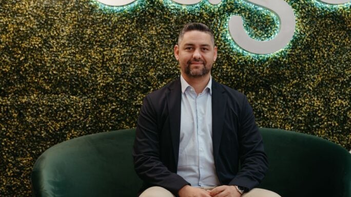 Una persona se sienta en un sofá verde frente a una pared con vegetación y letras iluminadas que forman la palabra Sage. Tres lámparas colgantes redondas cuelgan sobre ella. La persona lleva una chaqueta oscura, una camisa clara y pantalones color caqui.