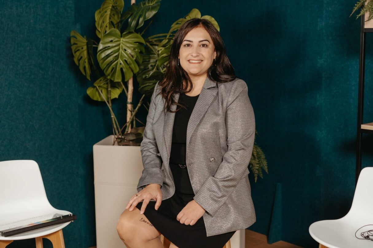 Una mujer de pelo largo y oscuro está sentada sonriendo en una habitación moderna. Lleva un blazer gris sobre un vestido negro. Detrás de ella hay una gran planta con hojas anchas y verdes. Cerca hay una silla blanca y una mesa pequeña. El fondo presenta una pared de color verde azulado intenso.