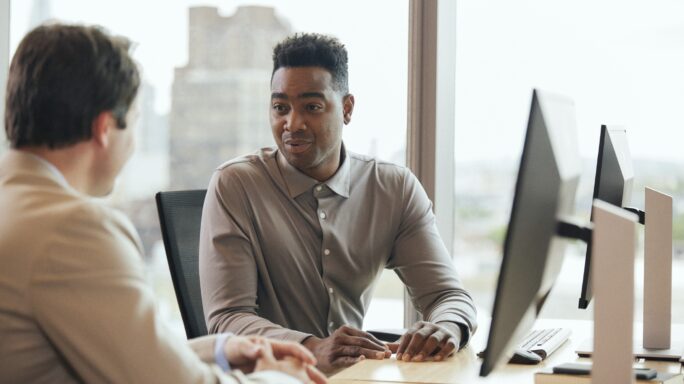 Deux hommes discutent, assis à leur bureau, devant une fenêtre. Il ya des écrans d'ordinateur sur les bureaux