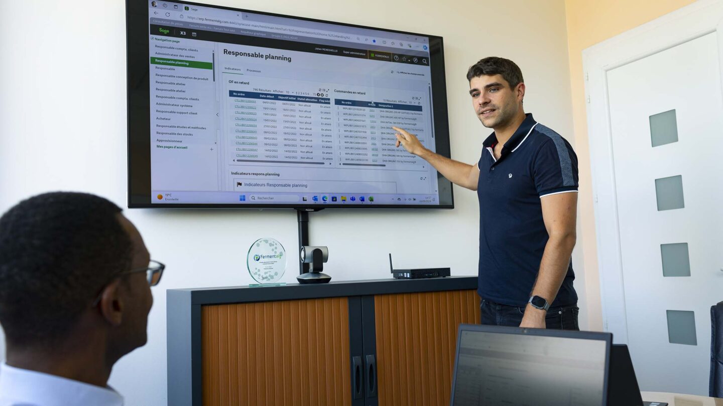 Dans une salle de réunion un homme présente un tableau de reporting sur grand écran à son collègue