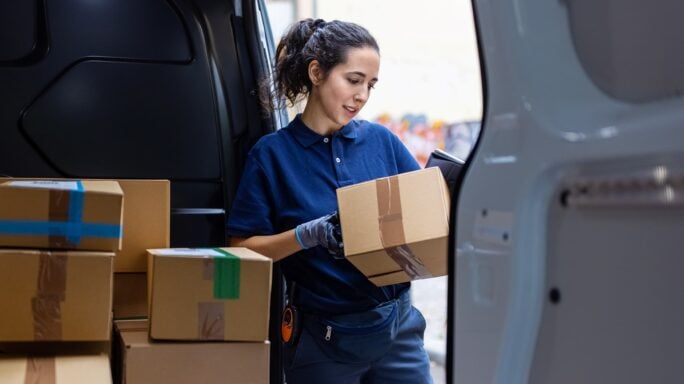 Uma entregadora usando um uniforme azul e luvas está parada na parte de trás de uma van, segurando um pacote. Vários outros pacotes estão empilhados ao redor dela dentro da van.