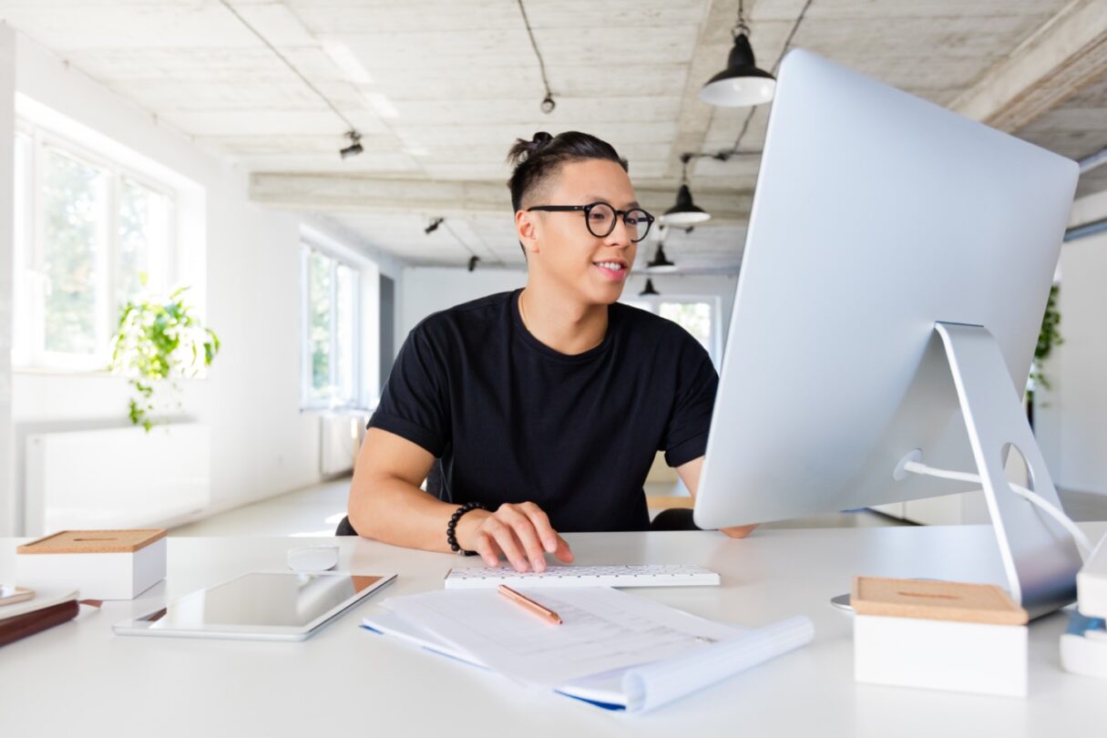 Um jovem sorridente trabalha no computador, sentado à secretária.