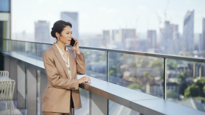 Uma mulher jovem, vestida com um blazer cinza e uma camisa branca, está sentada à frente de um computador portátil num ambiente de escritório moderno. Ela está a sorrir enquanto olha para o ecrã, sugerindo que está satisfeita com o que vê. Este cenário transmite uma atmosfera de produtividade e profissionalismo.